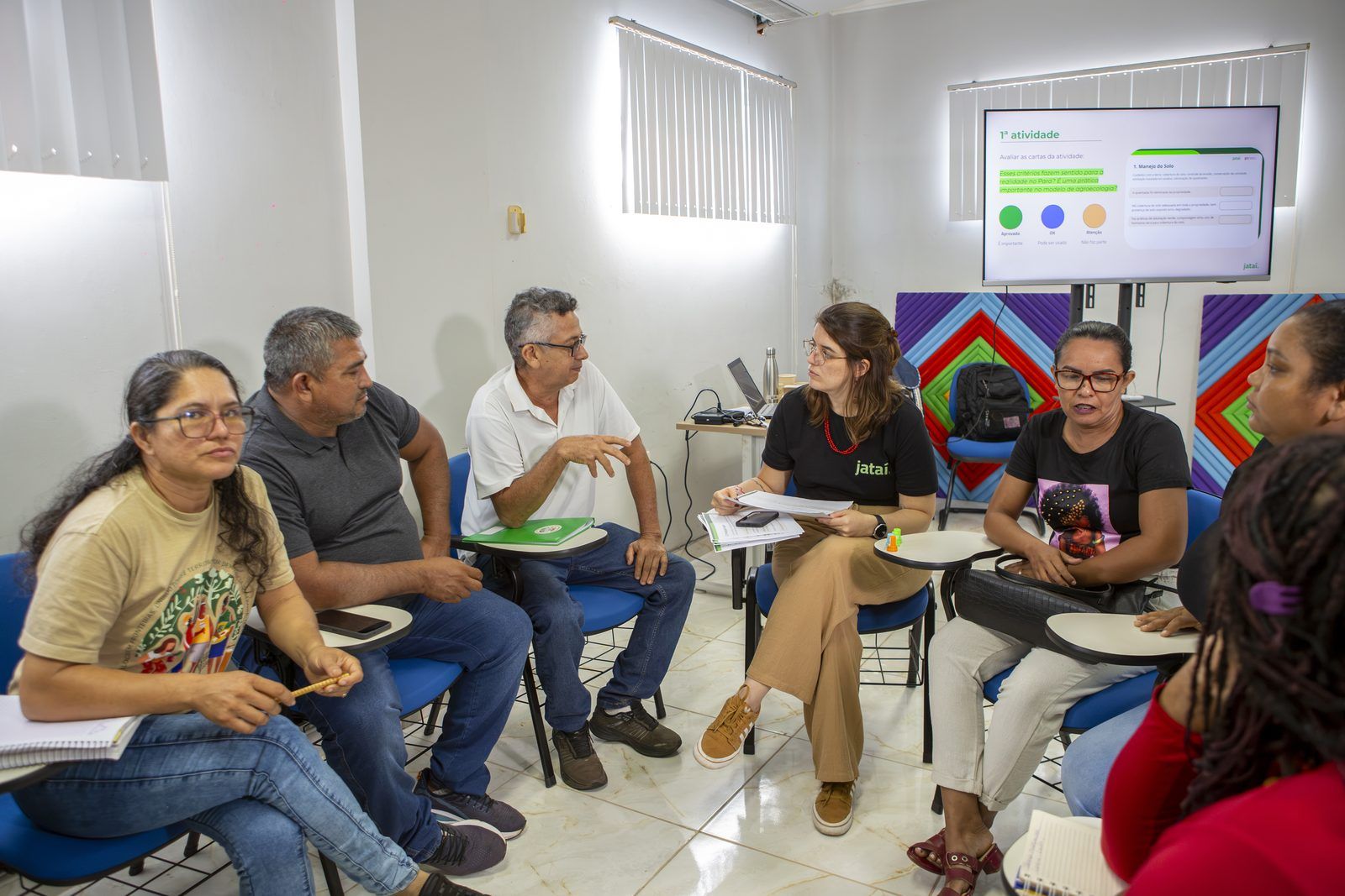 Integrante da equipe do Instituto Jataí conversa com cinco participantes sentados em roda, ao lado da TV com a primeira atividade da oficina.