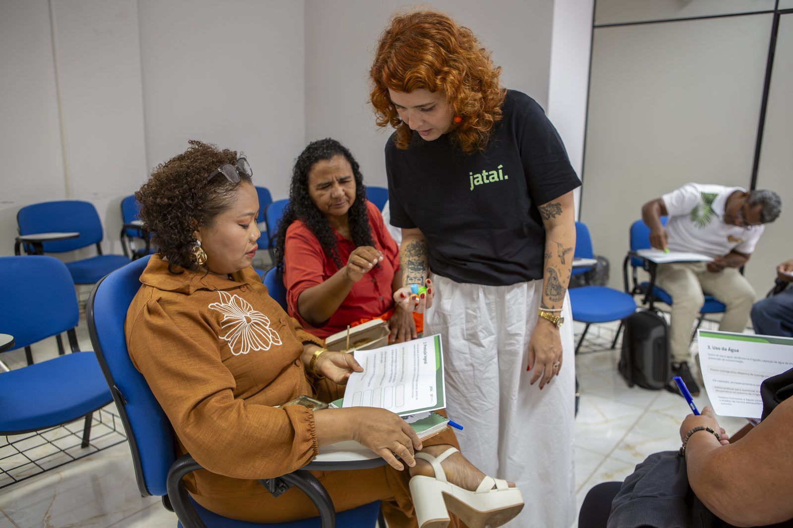 Facilitadora do Instituto Jataí mostra carimbos coloridos a duas participantes que leem uma ficha impressa.