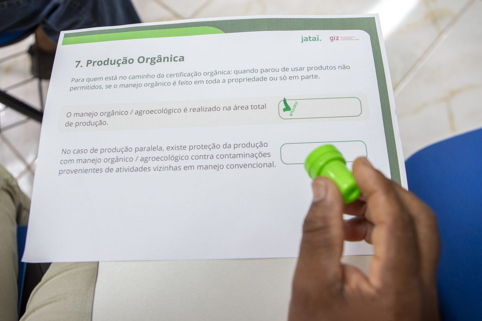 Mão segura um carimbo sobre a ficha “Produção Orgânica”, usada na atividade de diagnóstico.