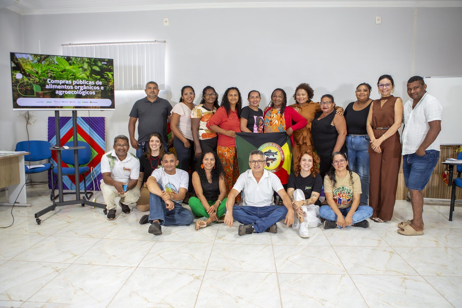 Participantes e equipe do Instituto Jataí posam juntos na sala da oficina, ao lado da TV com o slide “Compras públicas de alimentos orgânicos e agroecológicos”.